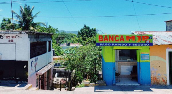 Between two run down concrete buildings steps lead down to a path with scraggly trees. The building on the left has a window covered with bars and in the basement is a hair salon. The building on the left is brightly colored with a sign reading Banca MJP, Pago Rapido Y Seguro. It has two chairs inside. At the top of the picture power lines run above the buildings and connect them with the background of a clear blue sky with a single wispy white cloud.